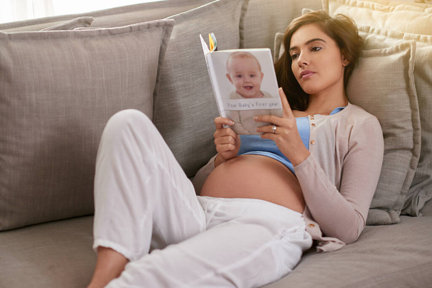 Vorbereitung auf die Reise durch die Mutterschaft. Aufnahme einer jungen Schwangeren, die zu Hause ein Babybuch liest. - Foto, Bild
