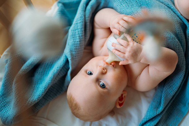 赤ん坊は哺乳瓶から牛乳を飲む。青い毛布の上に横たわる. - 写真・画像