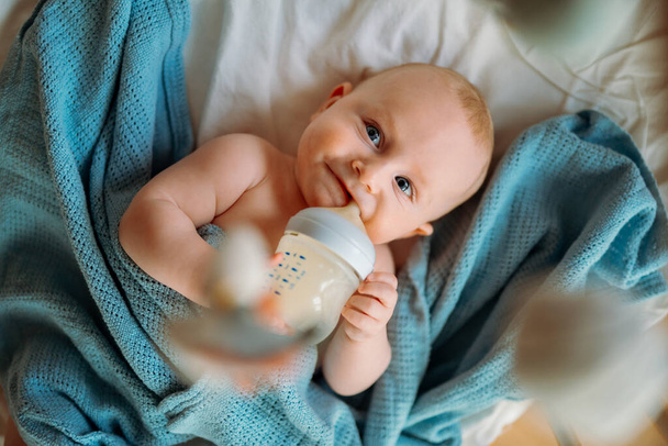 赤ん坊は哺乳瓶から牛乳を飲む。青い毛布の上に横たわる. - 写真・画像