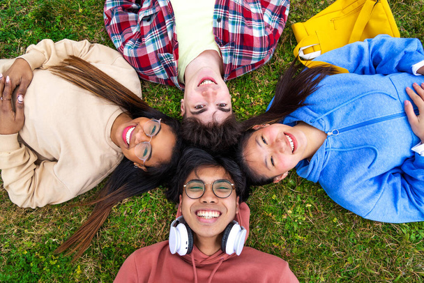 Gruppe fröhlicher und lächelnder multiethnischer College-Studenten, die im Gras liegen und mit Köpfen im Kreis in die Kamera schauen. - Foto, Bild