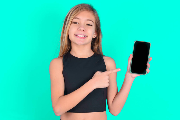 Посміхаючись блондинка маленька дівчинка в чорному спортивному одязі на зеленому тлі, показує і вказує на порожній екран телефону. Концепція реклами та комунікації. - Фото, зображення