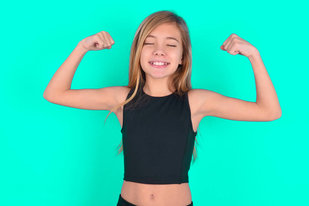 Sterke krachtige blonde kleine meisje dragen zwarte sport kleding over groene achtergrond met tanden glimlach, heft armen en toont biceps. Kijk naar mijn spieren.! - Foto, afbeelding