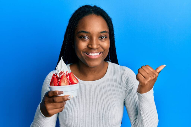 アフリカ系アメリカ人女性とともに編組髪食べるイチゴアイスポインティング親指上へザ側笑顔幸せとともにオープン口  - 写真・画像