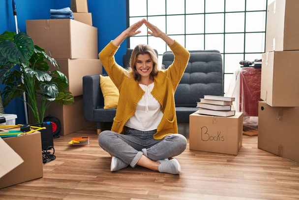 Young blonde woman smiling confident doing house gesture with hands at new home - 写真・画像