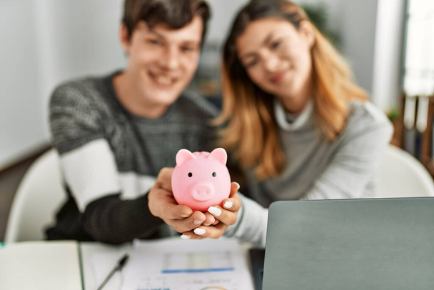 Young caucasian couple smiling happy holding piggy bank sitting on the table at home. - Фото, изображение