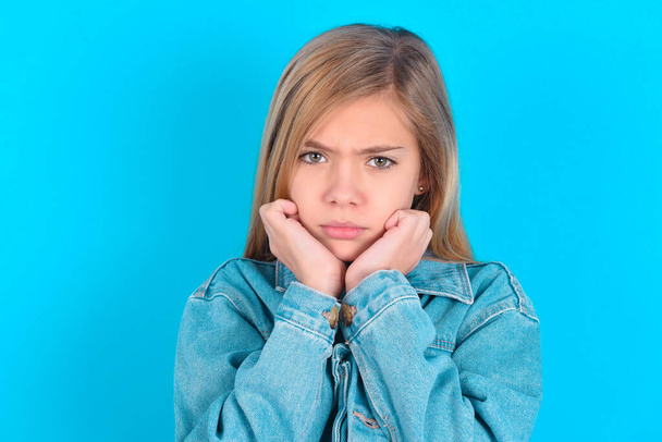 Portret smutnej blondynki dziewczynki w dżinsowej kurtce na niebieskim tle z twarzą w dłoniach - Zdjęcie, obraz