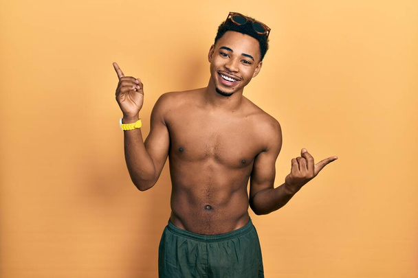 Jonge Afro-Amerikaanse man draagt zwemkleding en een zwembril en glimlacht vol vertrouwen wijzend met vingers naar verschillende richtingen. kopieerruimte voor reclame  - Foto, afbeelding