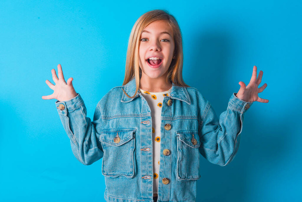 Delighted positieve blonde kleine jongen meisje dragen denim jas over blauwe achtergrond opent mond en armen palmen tot na het hebben van geweldig resultaat - Foto, afbeelding