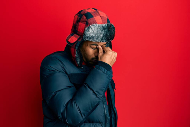 Young african american man with beard wearing winter hat and coat tired rubbing nose and eyes feeling fatigue and headache. stress and frustration concept.  - Foto, afbeelding