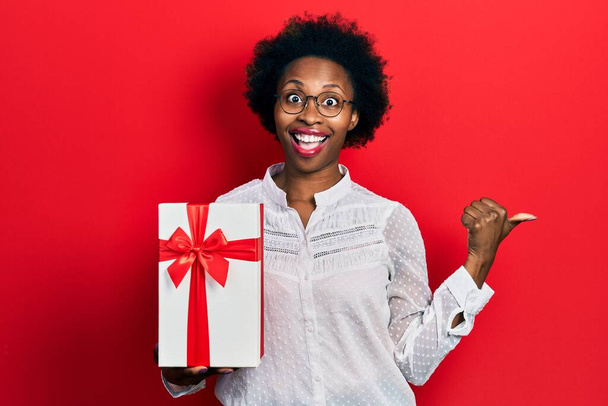 Young african american woman holding gift pointing thumb up to the side smiling happy with open mouth  - Photo, Image