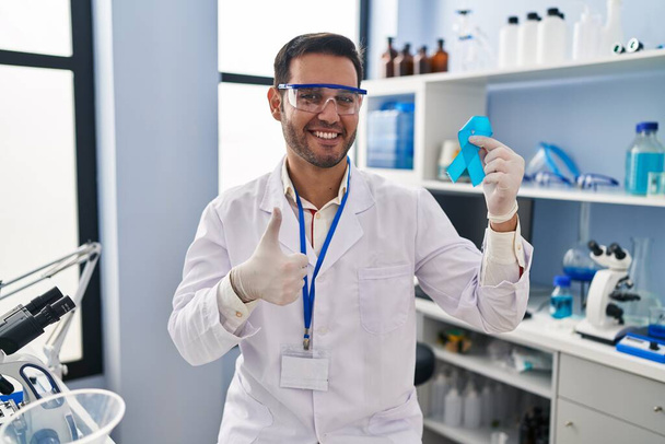 Mladý hispánec s vousy pracující v laboratoři vědec drží modrou stuhu s úsměvem šťastný a pozitivní, palec nahoru dělá vynikající a schvalovací znamení  - Fotografie, Obrázek