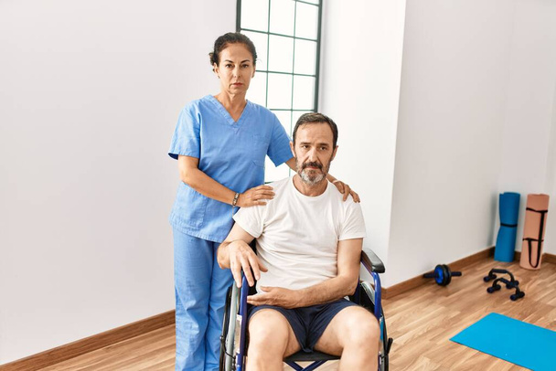 Homem hispânico de meia idade sentado em cadeira de rodas e enfermeira na clínica de reabilitação pensando atitude e expressão sóbria olhando auto confiante  - Foto, Imagem