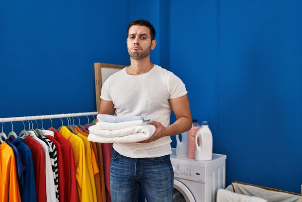 髭を生やした若いヒスパニック系の男性は、洗濯室で落ち込んできれいなタオルを折り畳んで保持し、苦痛を心配し、怒っていると恐れて泣いている。悲しい表情.  - 写真・画像
