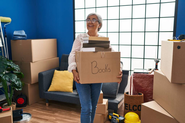 Keski-ikäinen nainen hymyilee luottavainen tilalla kirjoja paketti uudessa kodissa - Valokuva, kuva
