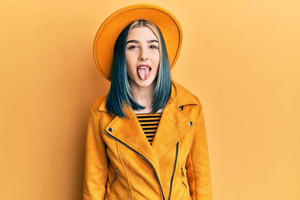 Junges modernes Mädchen mit gelbem Hut und Lederjacke, die die Zunge herausstreckt, mit lustiger Miene. Emotionales Konzept.  - Foto, Bild