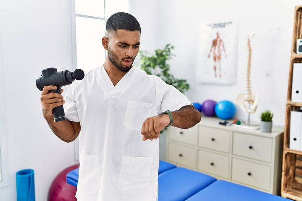 Young indian physiotherapist holding therapy massage gun at wellness center looking at the watch time worried, afraid of getting late  - Photo, Image