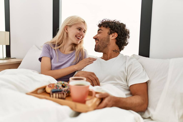 Νεαρό όμορφο ζευγάρι χαμογελά ευτυχισμένη έχοντας πρωινό στο κρεβάτι στο σπίτι. - Φωτογραφία, εικόνα