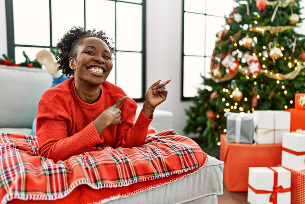 Fiatal afro-amerikai nő fekszik a kanapén karácsonyfa mosolyog, és a kamerába néz, két kézzel és ujjal az oldalán.  - Fotó, kép
