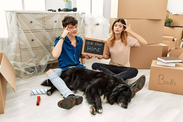 Mladý bělošský pár se psem drží naši první domácí tabuli v novém domě s úsměvem ukazuje na hlavu jedním prstem, skvělý nápad nebo myšlenka, dobrá paměť  - Fotografie, Obrázek