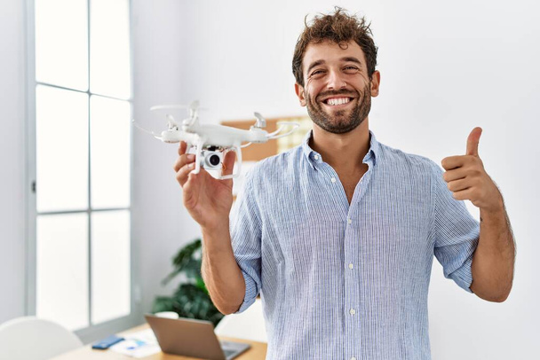 Νεαρός όμορφος άντρας που χρησιμοποιεί drone στο γραφείο αρχιτέκτονα χαμογελώντας χαρούμενος και θετικός, κάνει εξαιρετική δουλειά και σήμα έγκρισης  - Φωτογραφία, εικόνα