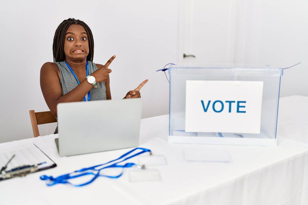 Jeune femme afro-américaine travaillant aux élections politiques assis par bulletin de vote pointant de côté inquiet et nerveux des deux mains, préoccupé et expression surprise  - Photo, image