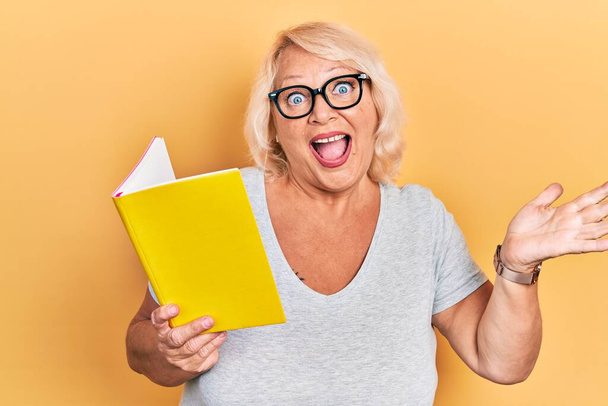 Middle age blonde woman reading a book celebrating achievement with happy smile and winner expression with raised hand  - Fotó, kép