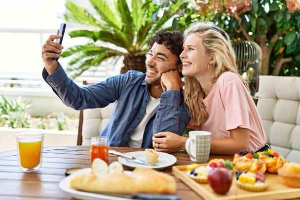 若いカップルが朝食を持っているし、テラスでテーブルの上に座ってスマートフォンで自撮り. - 写真・画像