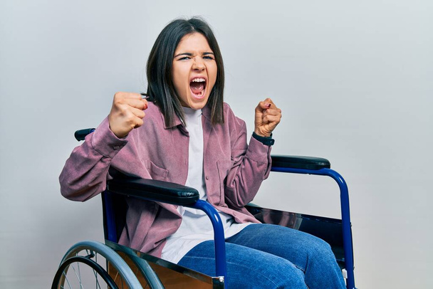 Genç esmer kadın kızgın ve kızgın bir şekilde tekerlekli sandalyede oturuyor öfkeyle bağırırken yumruklarını kaldırıyor. öfke ve agresif konsept.  - Fotoğraf, Görsel