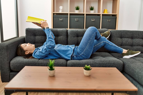 Młoda Chinka czyta książkę leżącą na kanapie w domu. - Zdjęcie, obraz