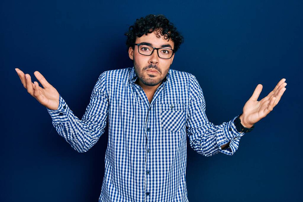 Junger hispanischer Mann in lässiger Kleidung und Brille ahnungslos und verwirrt mit offenen Armen, keine Ahnung und zweifelhaftem Gesicht.  - Foto, Bild