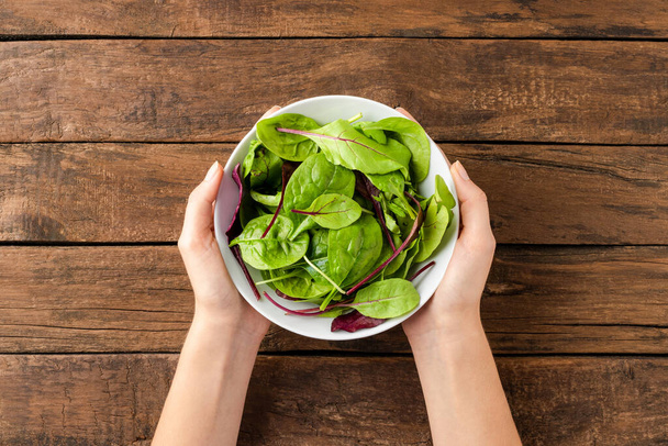 Overhead shot of woman hands holding green salad in bowl on rustic wooden background. Close up - Photo, image