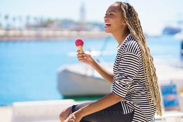 Zwarte vrouw die een aardbeienijs eet terwijl ze geniet van het uitzicht op de zeehaven. - Foto, afbeelding