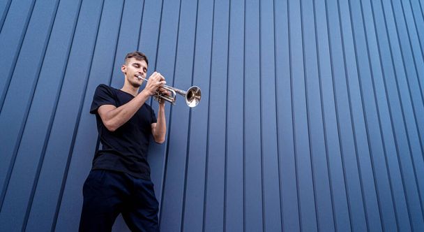 Büyük mavi duvarın yanında trompet çalan genç bir sokak müzisyeni. - Fotoğraf, Görsel