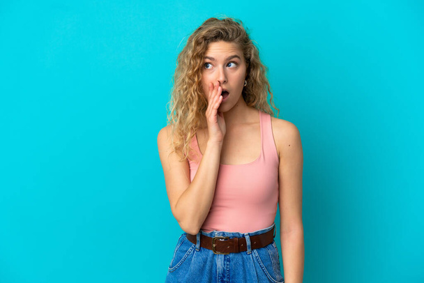 Young blonde woman isolated on blue background whispering something with surprise gesture while looking to the side - Фото, зображення