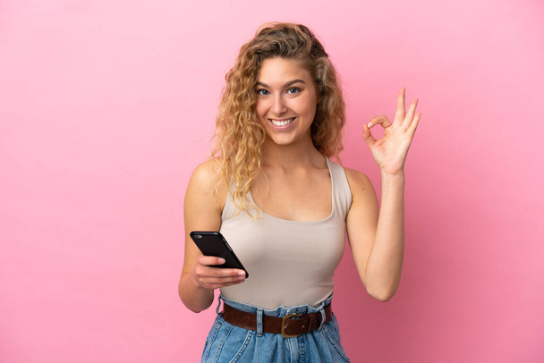 Young blonde woman isolated on pink background using mobile phone and doing OK sign - Foto, Bild