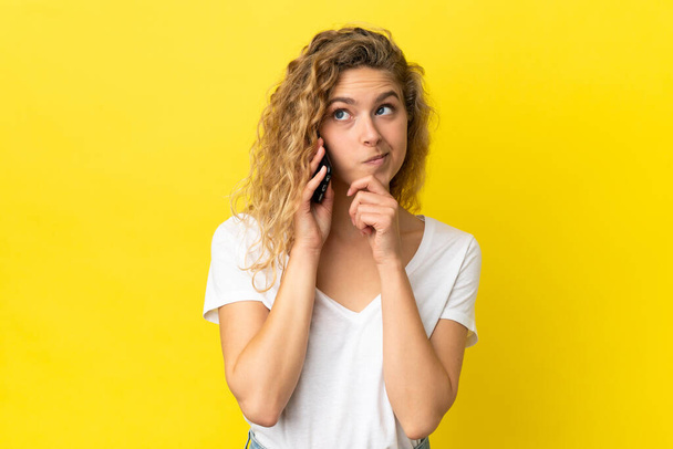 Young blonde woman using mobile phone isolated on yellow background and looking up - Foto, Bild