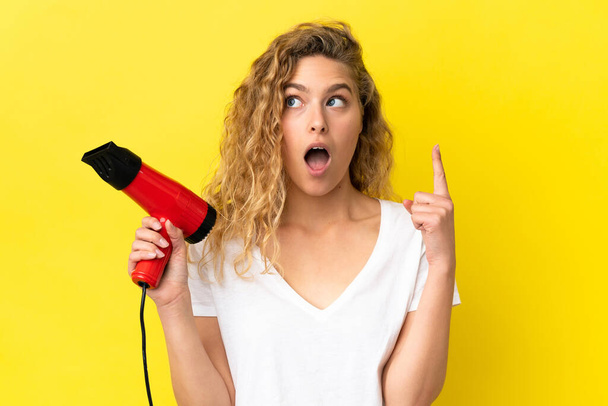 Young blonde woman holding a hairdryer isolated on yellow background thinking an idea pointing the finger up - Fotoğraf, Görsel