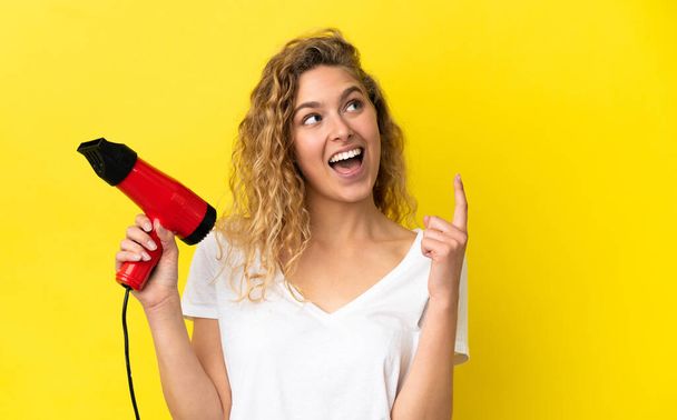 Young blonde woman holding a hairdryer isolated on yellow background pointing up a great idea - Foto, afbeelding
