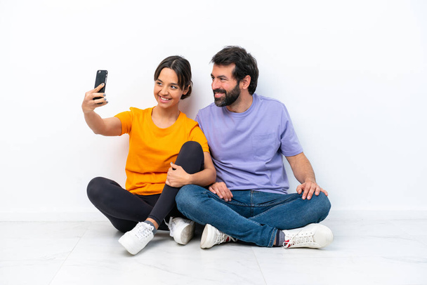 Young caucasian couple sitting on the floor isolated on white background making a selfie with the mobile - 写真・画像