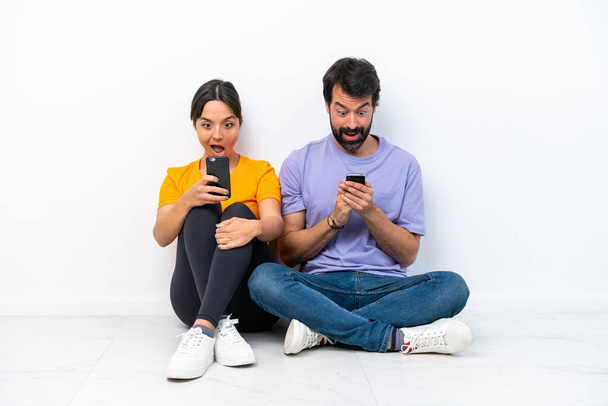Young caucasian couple sitting on the floor isolated on white background Surprised and sending a message or email with the mobile - Photo, Image