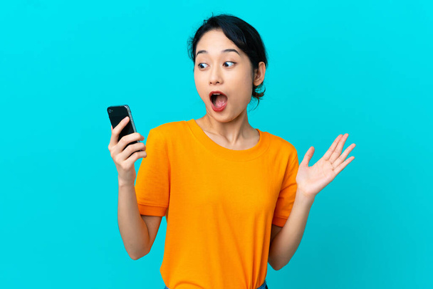 Young Vietnamese woman isolated on blue background looking at the camera while using the mobile with surprised expression - 写真・画像