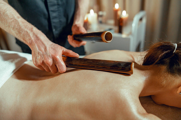 Мужчина-терапевт делает массаж спины женщине, используя полые бамбуковые трости - Фото, изображение