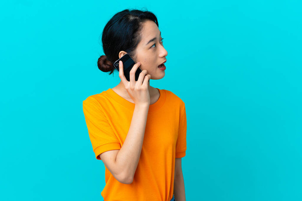 Jeune femme vietnamienne isolée sur fond bleu gardant une conversation avec le téléphone portable avec quelqu'un - Photo, image