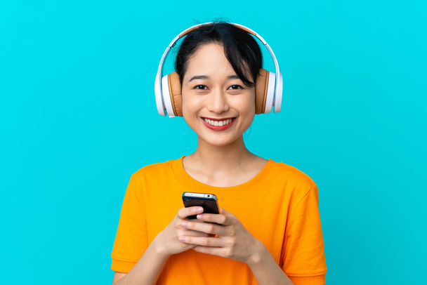 Junge Vietnamesin isoliert auf blauem Hintergrund Musik hören mit einem Handy und Blick nach vorne - Foto, Bild