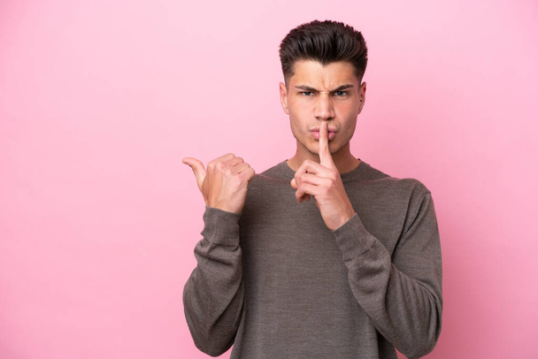 Young caucasian man isolated on pink background pointing to the side and doing silence gesture - Photo, Image