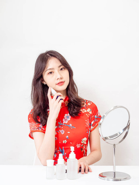 Portrait of a young asian woman in traditional cheongsam qipao dress looks at mirror and does skin care isolated on white background. - Fotografie, Obrázek