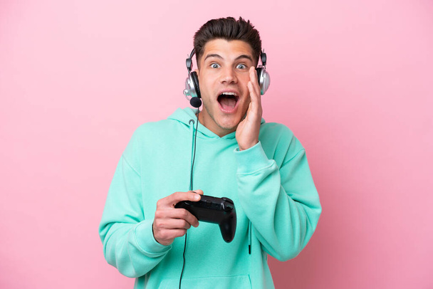 Giovane uomo caucasico bello che gioca con un controller di videogiochi isolato su sfondo rosa con sorpresa e espressione facciale scioccata - Foto, immagini