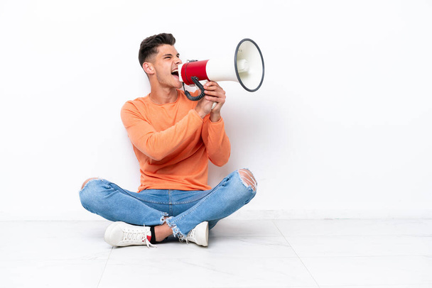 Young man sitting on the floor isolated on white background shouting through a megaphone - Photo, image
