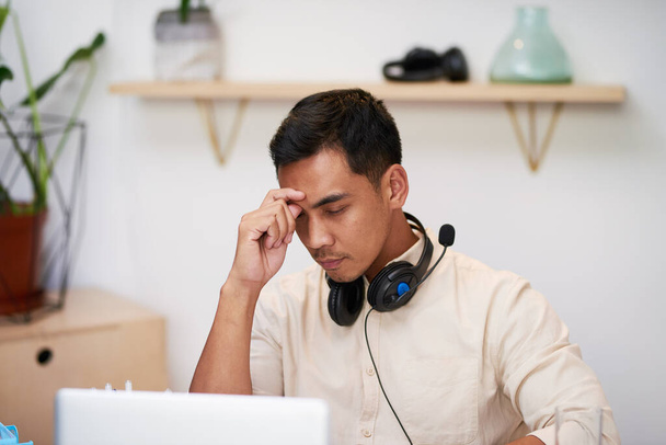 Un joven hombre de negocios asiático se frota los ojos en su escritorio en la oficina buscando molesto - Foto, Imagen