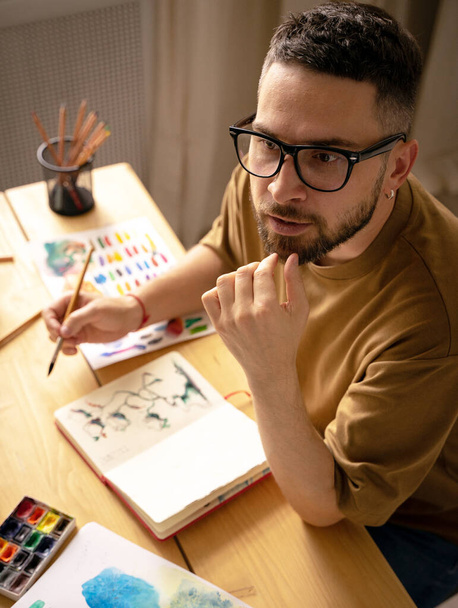 Egy fiatal művész rajzol egy ecsettel az asztalán. Egy kreatív ember portréja a műtermében. Kreatív stúdió, életmód, egy műalkotás létrehozásának folyamata, az inspiráció keresése. - Fotó, kép